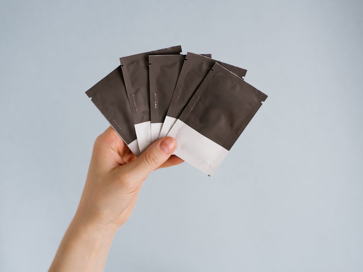 Hand holding a fan of small blank foil sachets against a pale blue wall